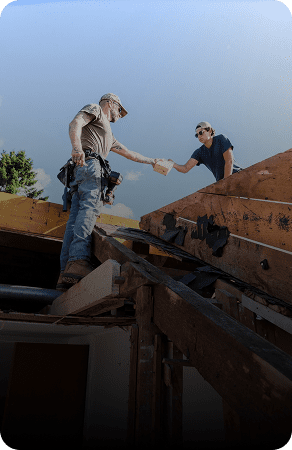 Two construction workers passing a tool on a roof under clear skies.